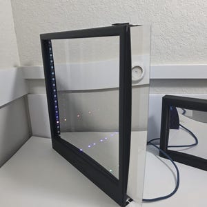 May include: A black framed LED light panel with a clear glass surface. The panel has a row of multi-coloured lights along one edge. A second, similar panel is visible in the background.