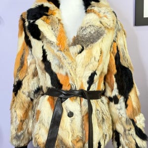 May include: A multi-coloured fur coat with a black leather belt. The coat features a mix of black, orange, and cream-coloured fur. The coat is displayed on a mannequin. The belt is tied at the waist.
