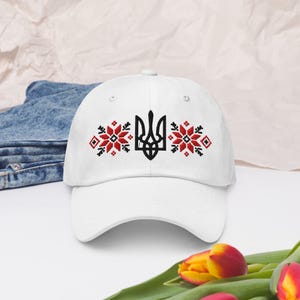 Puede incluir: Una gorra de béisbol blanca con un símbolo ucraniano Tryzub bordado en negro, centrado entre dos patrones florales geométricos en rojo y negro. La gorra cuenta con una corona estructurada, una visera curva y una correa ajustable.