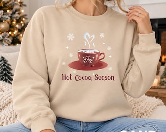 May include: A sand-coloured crewneck sweatshirt featuring a graphic of a dark red mug filled with hot cocoa and marshmallows. The mug has the word Joy written on it. Below the mug, the text reads Hot Cocoa Season, surrounded by white snowflakes.