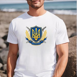 May include: A white short-sleeved t-shirt featuring a central graphic of a blue and gold shield with a yellow trident emblem. The shield is flanked by two crossed silver swords and golden wheat stalks, with a blue ribbon banner displaying 1991.