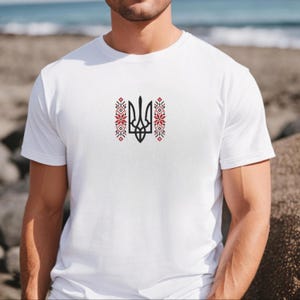 May include: A white short-sleeved t-shirt featuring a central black Ukrainian trident symbol, known as the Tryzub, flanked by two vertical decorative patterns in red and black geometric floral motifs centered on the chest area.