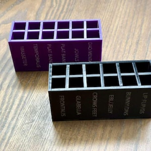 May include: Two rectangular organizers, one purple and one black, each with multiple compartments. The purple organizer has text that reads "MASSETER" and "TEMPORALIS." The black organizer has text that reads "FRONTALIS" and "GLABELLA."