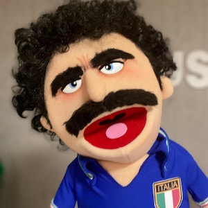 Może przedstawiać: Marionetka z szczegółową twarzą, kręconymi czarnymi włosami, dużymi czarnymi wąsami i niebieskimi oczami. Marionetka nosi niebieską koszulę z białą, zieloną i czerwoną odznaką z napisem "ITALIA". Usta marionetki są otwarte, odsłaniając czerwone wnętrze i różowy język.