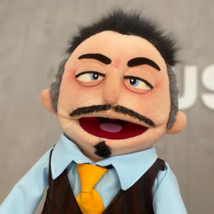 Może przedstawiać: Marionetka z ludzką twarzą, ubrana w jasnoniebieską koszulę z kołnierzykiem, brązową kamizelkę w pionowe paski i żółty krawat. Marionetka ma czarne wąsy, kozacką bródkę i ciemne włosy. Usta marionetki są otwarte, odsłaniając różowe wnętrze.