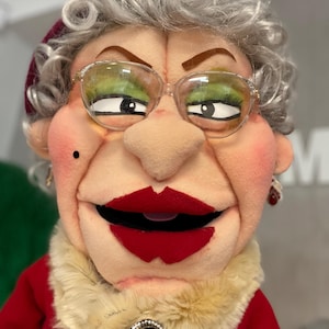 Może przedstawiać: Zbliżenie kukiełki z surowym wyrazem twarzy. Kukiełka ma siwe włosy, okulary i czerwoną szminkę. Nosi czerwony strój z kołnierzem z imitacji futra i broszką z kameą. Oczy kukiełki są zielone.