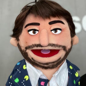 Może przedstawiać: Lalka z brązowymi włosami, brodą i czerwonymi ustami. Lalka ma na sobie granatową marynarkę z kolorowym wzorem Pac-Mana, białą koszulę z kołnierzykiem i pasujący krawat. Lalka ma duże, wyraziste oczy.