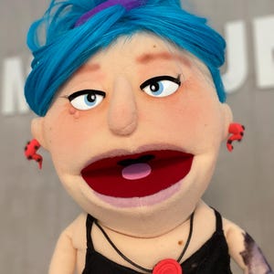 Może przedstawiać: Lalka z niebieskimi włosami, niebieskimi oczami i czerwonymi kolczykami. Lalka ma na sobie czarny top i czerwoną zawieszkę w kształcie róży. Usta lalki są otwarte, odsłaniając czerwone wnętrze. Lalka ma tatuaż na ramieniu.