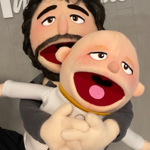 Może przedstawiać: Para ręcznie robionych kukiełek. Jedna kukiełka to brodaty mężczyzna z ciemnymi włosami i szarą koszulą. Druga kukiełka to dziecko z żółtym kołnierzem i koszulą z napisem "KEEPER". Obie kukiełki mają otwarte usta.