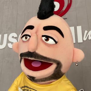 Może przedstawiać: Lalka z czarnym irokezem i żółtą koszulką. Lalka ma brodę i mały kolczyk. Na koszulce widnieje napis "OFFICIAL FAN 461M". Usta lalki są otwarte.