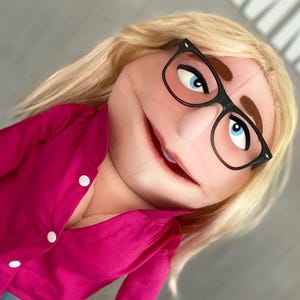 Może przedstawiać: Lalka z blond włosami, niebieskimi oczami i okularami w czarnych oprawkach. Lalka ma na sobie jasnoróżową koszulę zapinaną na guziki z białymi guzikami. Usta lalki są lekko otwarte, co sugeruje przyjazny wyraz twarzy.