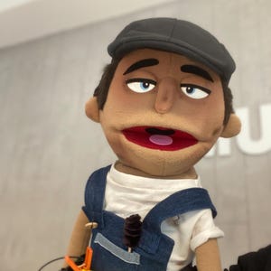 Może przedstawiać: Ręcznie wykonana kukiełka przedstawiająca mężczyznę o brązowej skórze, niebieskich oczach i czapce. Ma na sobie białą koszulę i niebieskie dżinsowe ogrodniczki. Usta kukiełki są otwarte, ukazując różowy język.