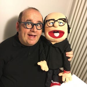 Może przedstawiać: Mężczyzna i kukiełka, obaj w okularach i czarnych koszulach, pozują do zdjęcia. Kukiełka ma duże otwarte usta i czerwone buty. Mężczyzna się uśmiecha, a kukiełka jest trzymana w jego ramionach.