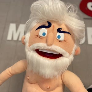 Może przedstawiać: Pluszowa kukiełka przedstawiająca starszego mężczyznę z białymi włosami i pasującą brodą. Kukiełka ma niebieskie oczy, szeroko otwarte usta i nie nosi ubrania. Ręce kukiełki są wyciągnięte, a tło jest rozmyte szare.