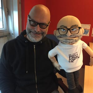 Puede incluir: Un hombre y una marioneta con gafas posan para una foto. El hombre lleva una sudadera negra con cremallera y gafas. La marioneta tiene una camiseta blanca con texto y pantalones grises. El fondo es una pared roja.