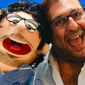 May include: A man and a puppet with glasses are smiling widely. The puppet has a beard and is wearing a blue shirt. The man is wearing a light blue button-down shirt. The background is a light blue and yellow.