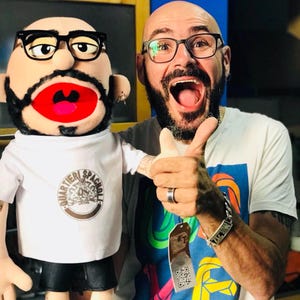 Może przedstawiać: Osoba z kukiełką. Kukiełka nosi okulary, białą koszulkę z okrągłym logo i czarne szorty. Osoba nosi okulary i białą koszulkę z kolorową grafiką. Oboje się uśmiechają.