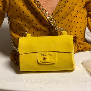 Może przedstawiać: Mała, jasnożółta torebka z pikowanym wzorem i złotym łańcuszkiem. Torebka posiada zapięcie na klapę z logo. Tło obejmuje żółty top w kropki.