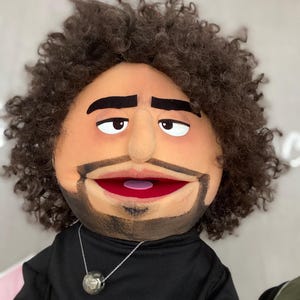 Może przedstawiać: Ręcznie wykonana kukiełka z opaloną twarzą, brązowymi kręconymi włosami i brodą. Kukiełka ma czarną koszulę i srebrny naszyjnik z okrągłym wisiorkiem. Usta kukiełki są otwarte, odsłaniając czerwone wnętrze.