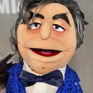 Może przedstawiać: Marionetka z opaloną twarzą, niebieskimi oczami i siwymi włosami. Marionetka ma na sobie niebieską kurtkę z cekinami, białą koszulę i granatową muszkę. Usta marionetki są otwarte, ukazując różowy język.