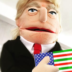 Może przedstawiać: Marionetka z blond włosami, niebieskimi oczami i czerwonymi ustami, ubrana w czarny garnitur, białą koszulę i czerwoną krawat. Marionetka trzyma małą flagę z gwiazdami i paskami po jednej stronie oraz czerwonymi, białymi i zielonymi paskami po drugiej.