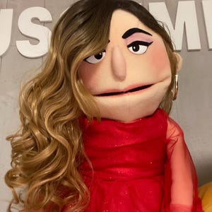 Może przedstawiać: Lalka z długimi, falowanymi blond włosami i czerwoną sukienką. Lalka ma neutralny wyraz twarzy z makijażem, w tym cienie do powiek i szminkę. Tło ma białe litery "US MI".