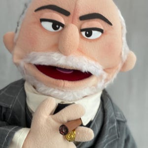 Może przedstawiać: Marionetka starszego mężczyzny z białymi wąsami i brwiami, ubranego w szary garnitur w paski, białą koszulę i czarny krawat. Marionetka trzyma cygaro i ma na palcu dwa złote pierścienie.