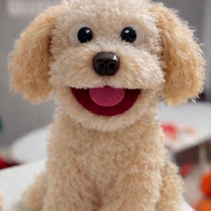 May include: A plush, light tan dog puppet with large, black eyes and a smiling red mouth. The puppet has fluffy fur and is sitting upright, showcasing its friendly expression. The dog's nose is a dark brown color.
