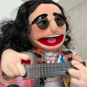 Może przedstawiać: Marionetka z długimi czarnymi włosami, okrągłymi okularami przeciwsłonecznymi i czerwonymi ustami, grająca na małej gitarze akustycznej. Marionetka ma na sobie kamizelkę na białej koszulce z napisem "RE" i "LEGALIZE". Na kamizelce widoczne są naszywki.