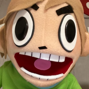 May include: A close-up of a plush mascot head with large, expressive eyes and a wide, open mouth. The mascot has tan hair, black eyebrows, and a green shirt. The mouth is red with white teeth and a pink tongue.
