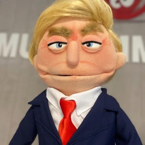 Może przedstawiać: Marionetka z blond peruką, niebieskimi oczami i surowym wyrazem twarzy. Marionetka ubrana jest w granatowy garnitur, białą koszulę i czerwoną krawat. Twarz marionetki jest w kolorze brzoskwiniowym.