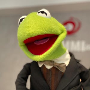 Puede incluir: Una marioneta de Kermit la Rana con una cara verde brillante, una boca roja grande y ojos negros. Lleva una chaqueta de traje negra, un chaleco marrón, una camisa blanca con cuello y una pajarita estampada. La marioneta está sonriendo.