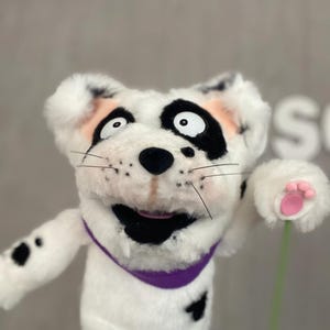Peut inclure: Marionnette de chien dalmatien en peluche, corps blanc, taches noires et collier violet. La marionnette a un visage souriant avec des marques noires autour des yeux et de la bouche, et des accents roses sur les oreilles et les coussinets.