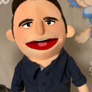 Może przedstawiać: Marionetka z opaloną twarzą, czarnymi włosami i granatową koszulą. Marionetka ma duże czerwone usta i uśmiecha się. Oczy marionetki są otwarte i patrzą w przód. Ujęcie z bliska marionetki.