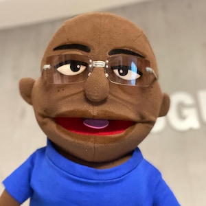 Może przedstawiać: Brązowa kukiełka w niebieskiej koszulce i okularach w przezroczystych oprawkach. Kukiełka ma szerokie usta z czerwonym wnętrzem i różowym językiem. Kukiełka ma ciemne brwi i małe uszy.