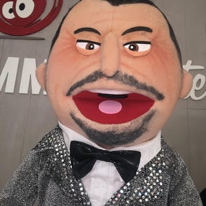 Może przedstawiać: Marionetka z opaloną twarzą, czarnymi włosami i wąsami. Nosi srebrną kurtkę z cekinami, białą koszulę i czarną muszkę. Usta marionetki są otwarte, ukazując różowy język.