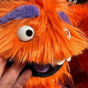 May include: Close-up of a fuzzy orange monster with large white eyes, purple accents, and a black mouth. The monster has white teeth and a purple tongue. The fur is long and shaggy, giving it a playful appearance.