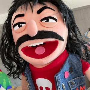 Może przedstawiać: Marionetka z beżową twarzą, czarnymi włosami i dużymi czarnymi wąsami. Nosi czerwoną koszulkę z białą grafiką czaszki i dżinsową kamizelkę. Usta marionetki są otwarte, odsłaniając czerwone wnętrze.