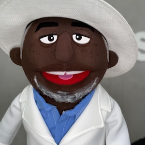 Może przedstawiać: Marionetka z ciemnobrązową twarzą, białymi włosami i białym kapeluszem. Marionetka ma na sobie białą kurtkę na niebieskiej koszuli. Marionetka ma szeroki uśmiech z czerwonymi ustami i białymi zębami.