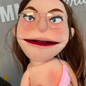 Może przedstawiać: Lalka z brązowymi włosami, w okularach w srebrnej oprawie i różowym topie od bikini. Lalka ma niebieskie oczy i czerwone usta. Tło zawiera litery "M" i "otte".