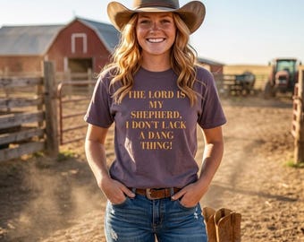 Può includere: Una persona indossa un cappello da cowboy marrone, una maglietta viola con la scritta "THE LORD IS MY SHEPHERD, I DON'T LACK A DANG THING!", jeans blu e stivali da cowboy marroni. Un fienile rosso e un trattore sono visibili sullo sfondo.