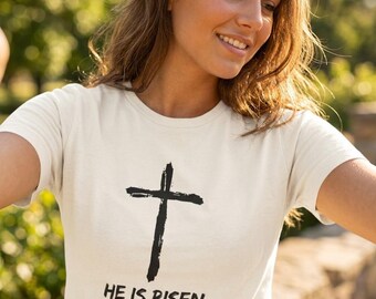 Può includere: T-shirt color crema con grafica a croce nera. Sotto la croce, il testo recita "HE IS RISEN" e "MATTEO 28:6" in nero. La maglietta ha maniche corte e girocollo.