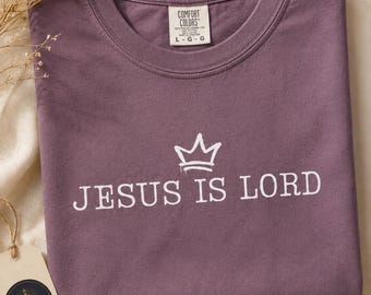 Puede incluir: Una camiseta de manga corta color ciruela con un gráfico blanco de una corona sobre el texto "JESUS IS LORD". La camiseta tiene cuello redondo y una etiqueta de la marca Comfort Colors en el cuello interior que indica la talla grande.