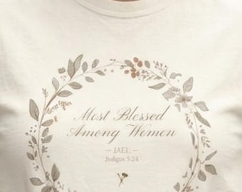 Può includere: T-shirt color crema con un design a ghirlanda floreale e la scritta "Most Blessed Among Women". La ghirlanda include foglie, fiori e bacche. Sotto il testo ci sono le parole "IAEL" e "Judges 5:24."