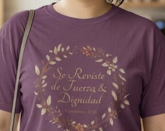 Può includere: Maglietta viola con un design a ghirlanda floreale dorata e la scritta "Se Reviste de Fuerza & Dignidad" e "Proverbios 31:25". La maglietta ha maniche corte e girocollo.