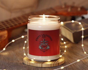 Può includere: Una candela accesa in un barattolo di vetro con un'etichetta rossa che recita "Happy Valentines Day" con un cuore, una croce e un design a rosa. La candela è appoggiata su un sottobicchiere rotondo di legno. Una chitarra e luci di fata sono sullo sfondo.