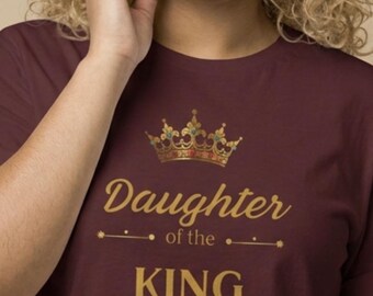 Può includere: T-shirt bordeaux con testo dorato e design a corona. La maglietta recita "Figlia del RE" con "Salmo 45:13" sotto. L'immagine mostra un primo piano della maglietta.