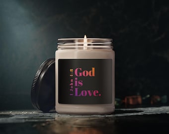 Può includere: Una candela accesa in un barattolo di vetro trasparente con coperchio nero. L'etichetta sul barattolo recita "Dio è Amore" in rosa e arancione, con il versetto biblico "1 Giovanni 4:8". La candela è su una superficie scura.