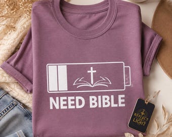Puede incluir: Una camiseta malva de manga corta con un gráfico blanco de un icono de batería baja que contiene una cruz y un libro abierto. Debajo del icono, el texto NEED BIBLE aparece en letras mayúsculas blancas. Una etiqueta negra indica REFLECT HIS LIGHT.