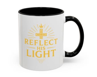 Può includere: Tazza in ceramica bianca con interno e manico neri. La tazza presenta la scritta "REFLECT HIS LIGHT" in oro, insieme a un disegno a croce e corona. La tazza è progettata per bevande calde.